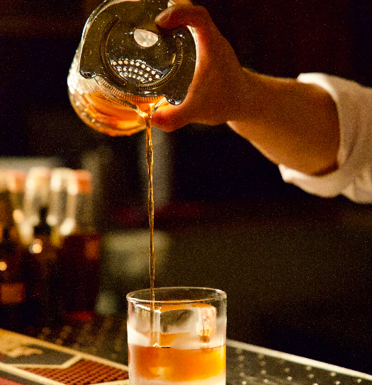 A bartender straining an amber cocktail from a shaker into a rocks glass over ice in a dimly lit bar.