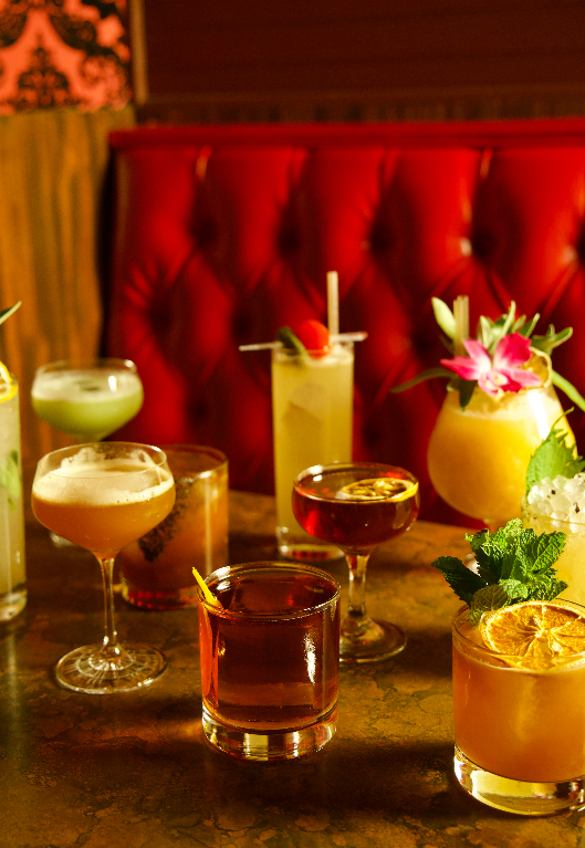 Assorted colorful cocktails arranged on a table against red tufted booth seating in a dimly lit bar setting