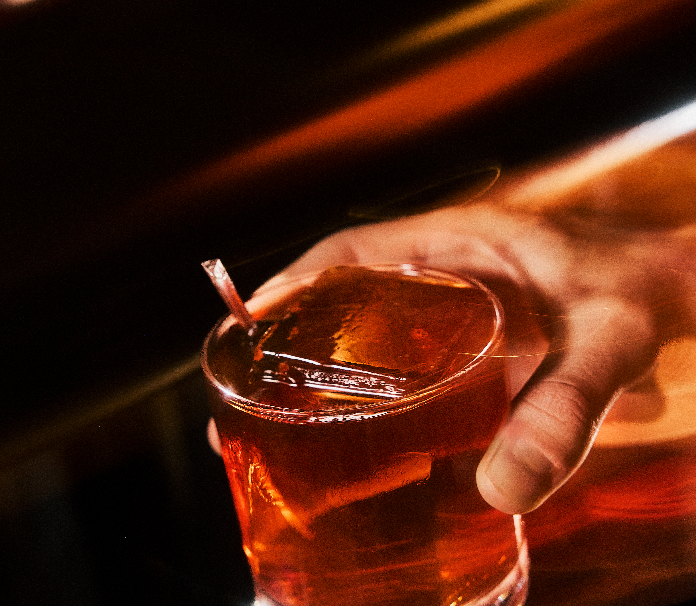 A hand holding a rocks glass with a deep amber cocktail over a large ice cube, glowing with warm red and orange tones against a dark background.