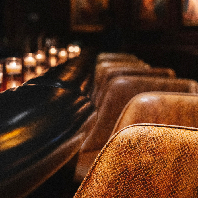 A dimly lit upscale bar or lounge interior featuring a row of tan leather chairs with a textured crocodile pattern alongside a dark leather banquette, with candles glowing softly in the background.