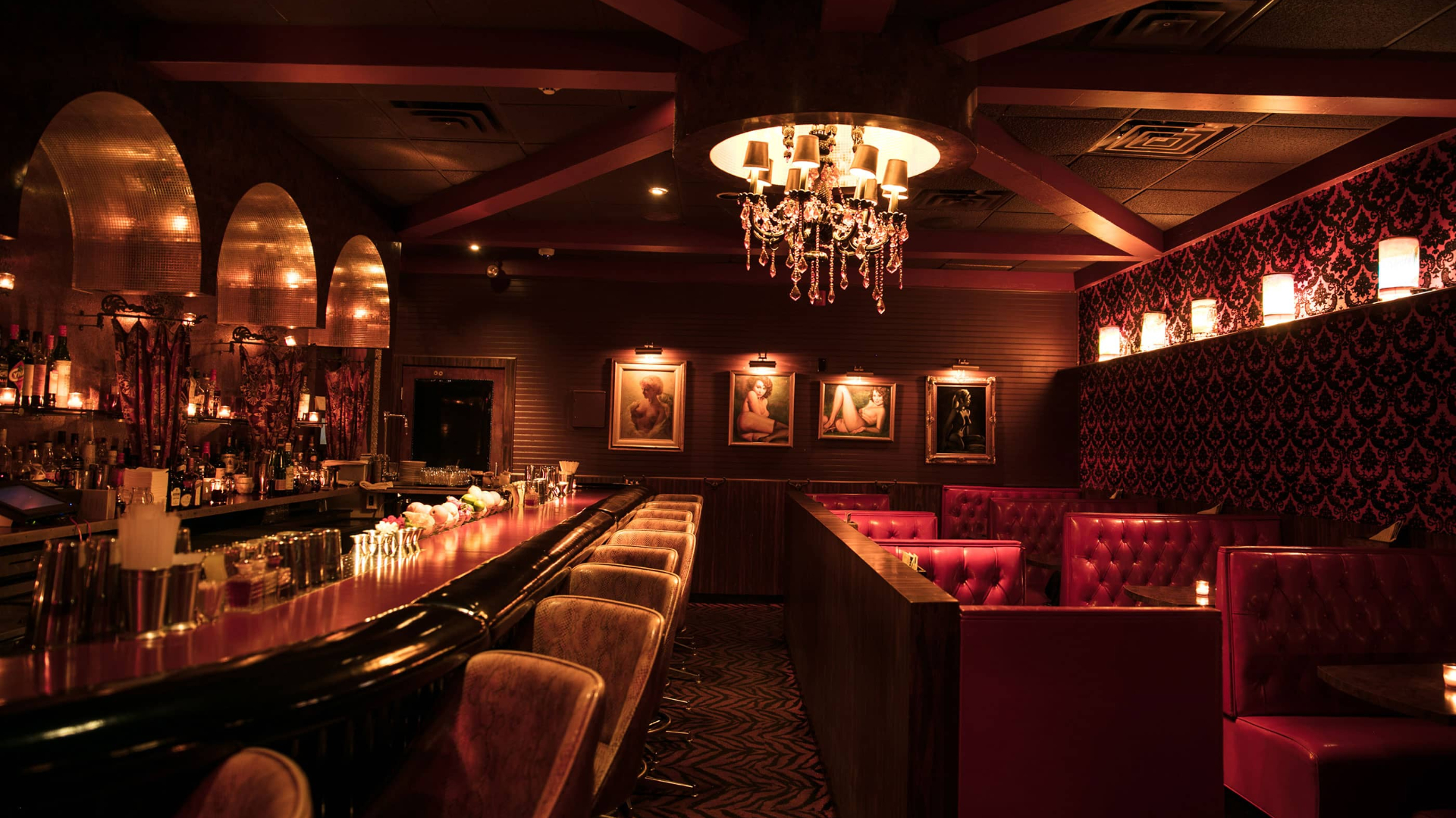 Upscale lounge interior with red tufted booths, crystal chandelier, illuminated bar, and vintage portraits on dark walls.