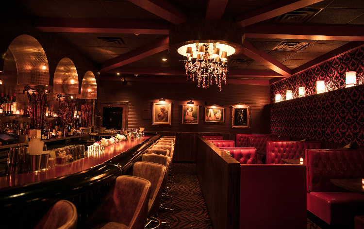 Upscale lounge interior with red tufted booths, crystal chandelier, illuminated bar, and vintage portraits on dark walls.